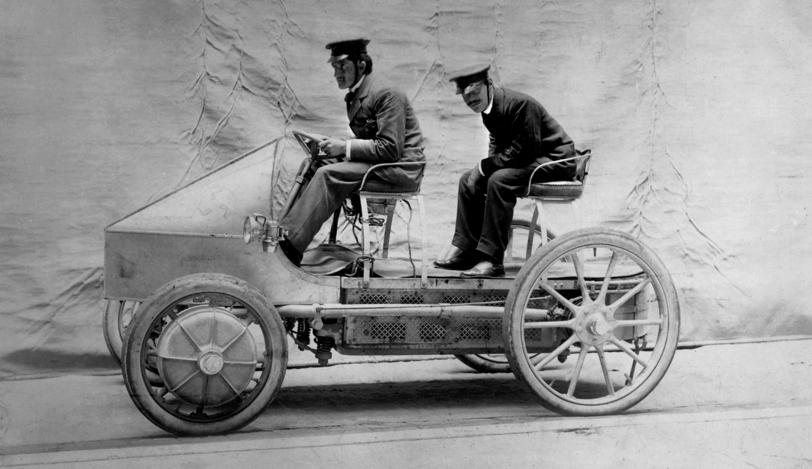 Two men are sitting in an early automobile on a studio background, both wearing hats and dressed in early 20th-century clothing.