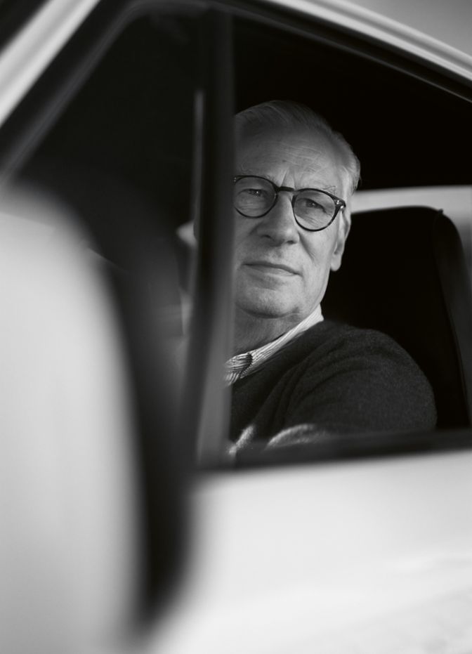 A man with glasses is seated inside a car, gazing out the window, with a soft light illuminating his face.
