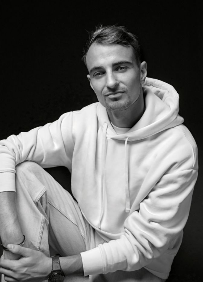 Ein junger Mann in hellem Hoodie sitzt vor dunklem Hintergrund und blickt entspannt in die Kamera.