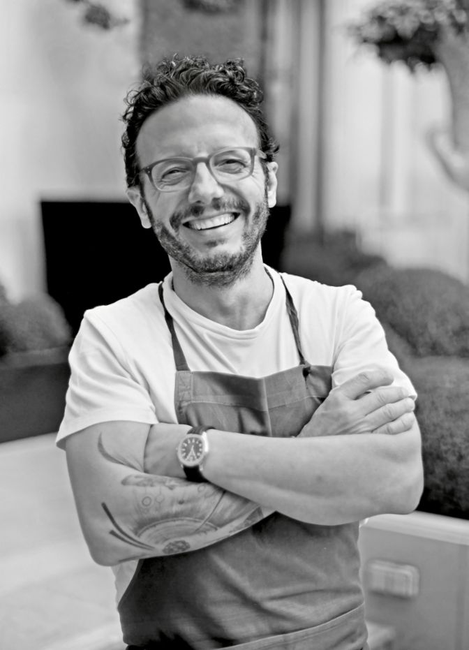 A smiling man with a tattoo on his arm is wearing a white t-shirt and an apron. 