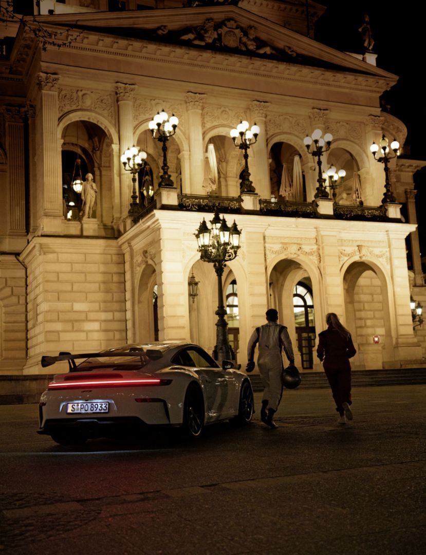 Zwei Personen in Rennanzügen laufen bei Nacht von einem Porsche 911 aus auf ein beleuchtetes Portal zu. 