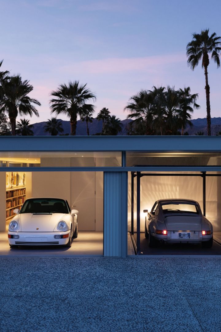 Two classic Porsches stand in a modern, illuminated garage building, surrounded by palm trees and at dusk.