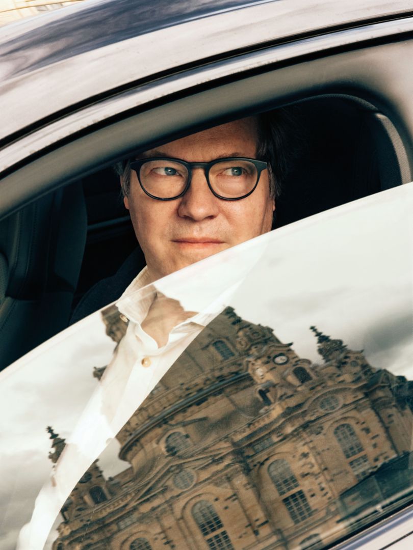 A man wearing glasses looks thoughtfully out of a car window, while a historic building is reflected in the window pane.