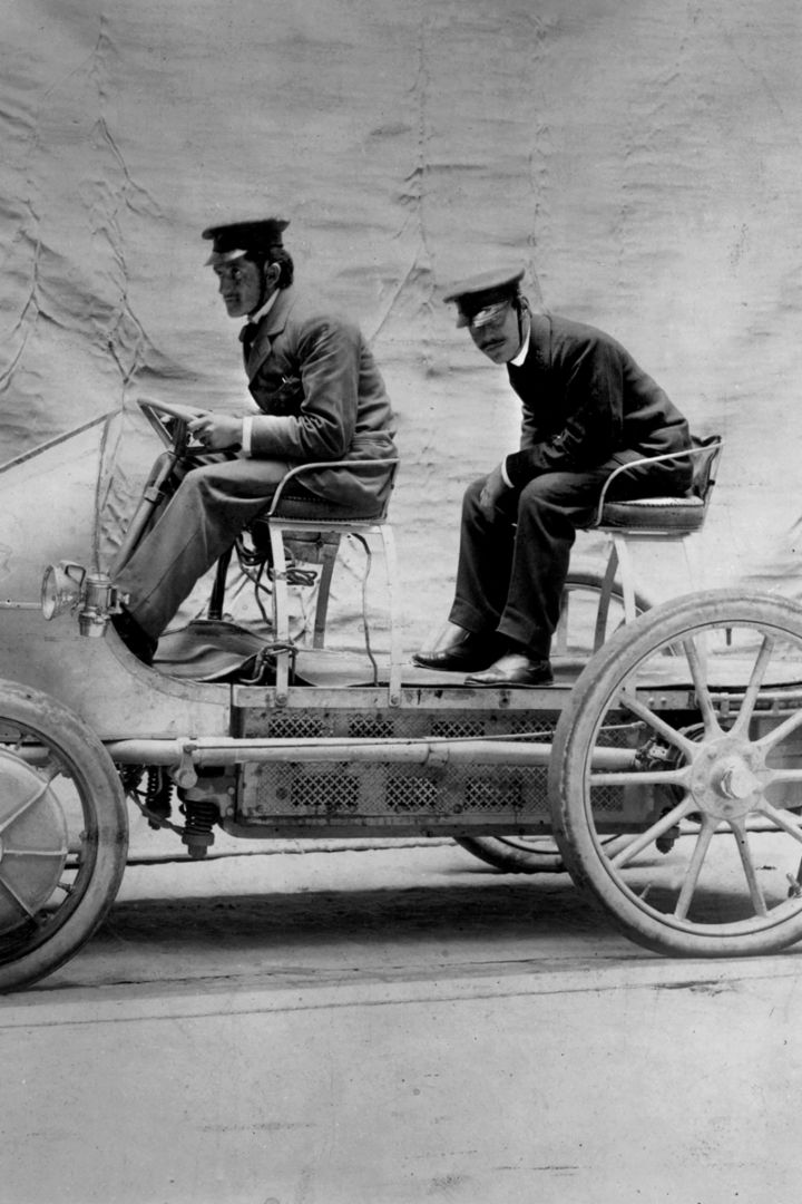 Two men are sitting in an early automobile on a studio background, both wearing hats and dressed in early 20th-century clothing.