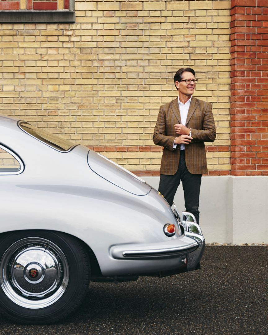 A man in a brown blazer stands next to a silver Porsche 356 and smiles in front of a brick wall.