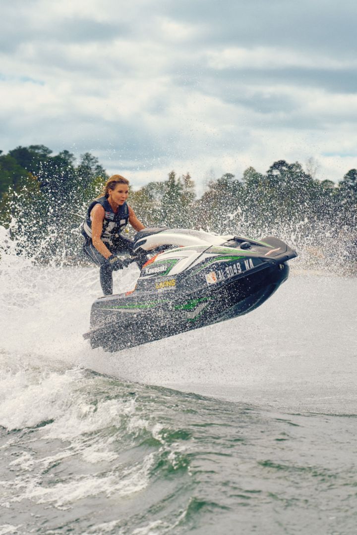 Eine Frau springt mit einem Jetski über eine Wasserwelle – im Hintergrund sind am Ufer Häuser und Bäume zu sehen. 