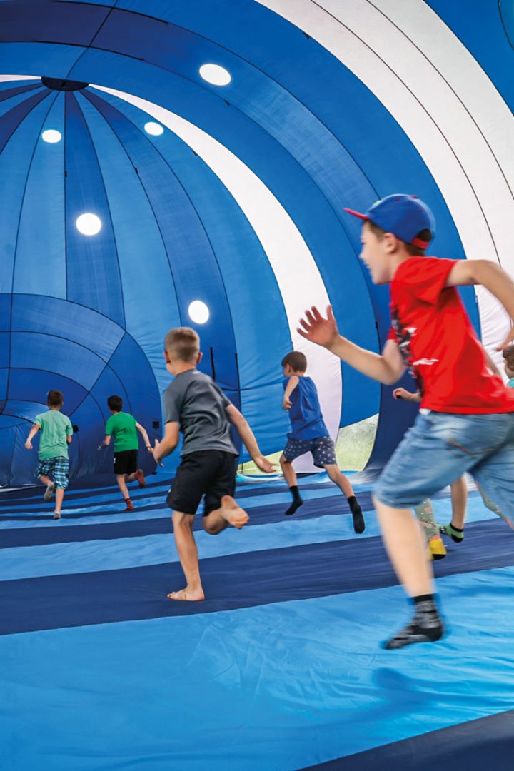 Kinder rennen barfuß und lachend in einer blauen, zeltartigen Struktur, die aussieht wie ein großes, aufblasbares Tunnelspielgerät. 