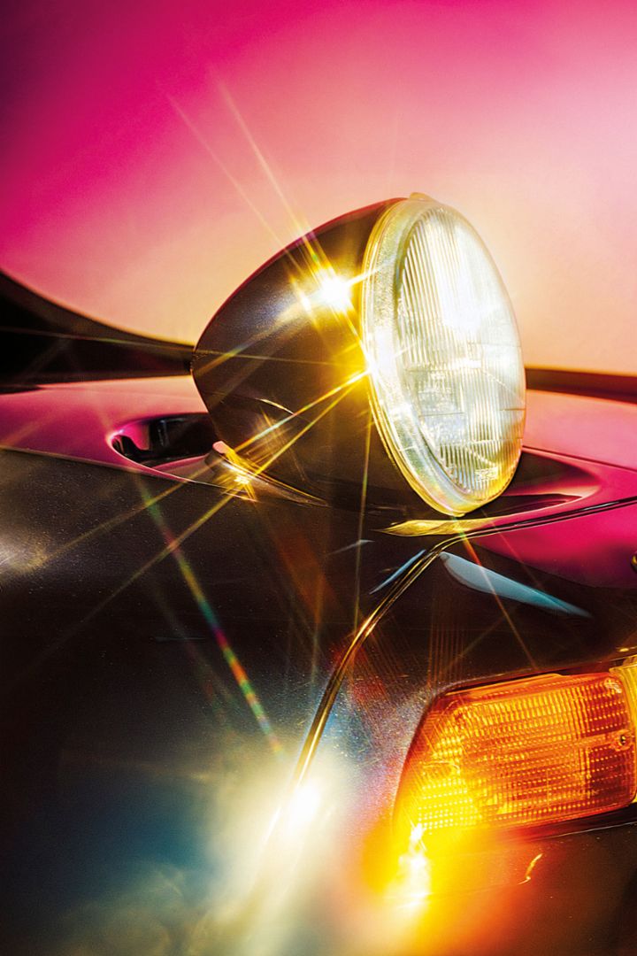 Eine Nahaufnahme des Scheinwerfers eines Porsche, der im Sonnenlicht glänzt. Das Licht reflektiert auf dem metallischen Lack und erzeugt eine auffällige Leuchterscheinung.