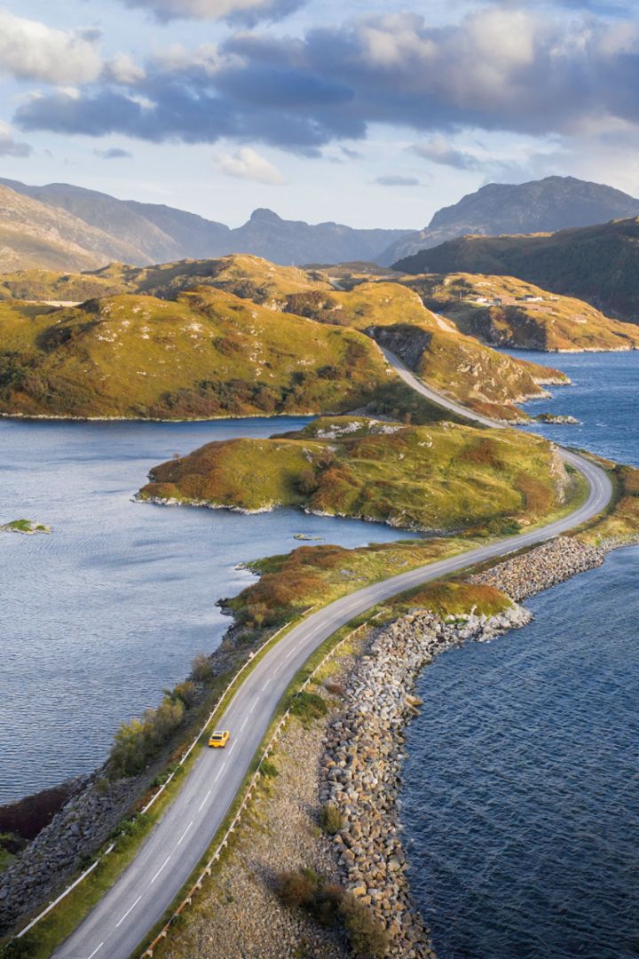Eine malerische Küstenstraße führt entlang eines Gewässers, umgeben von Hügeln und Gebirgen, mit einem gelben Porsche, der darauf fährt.