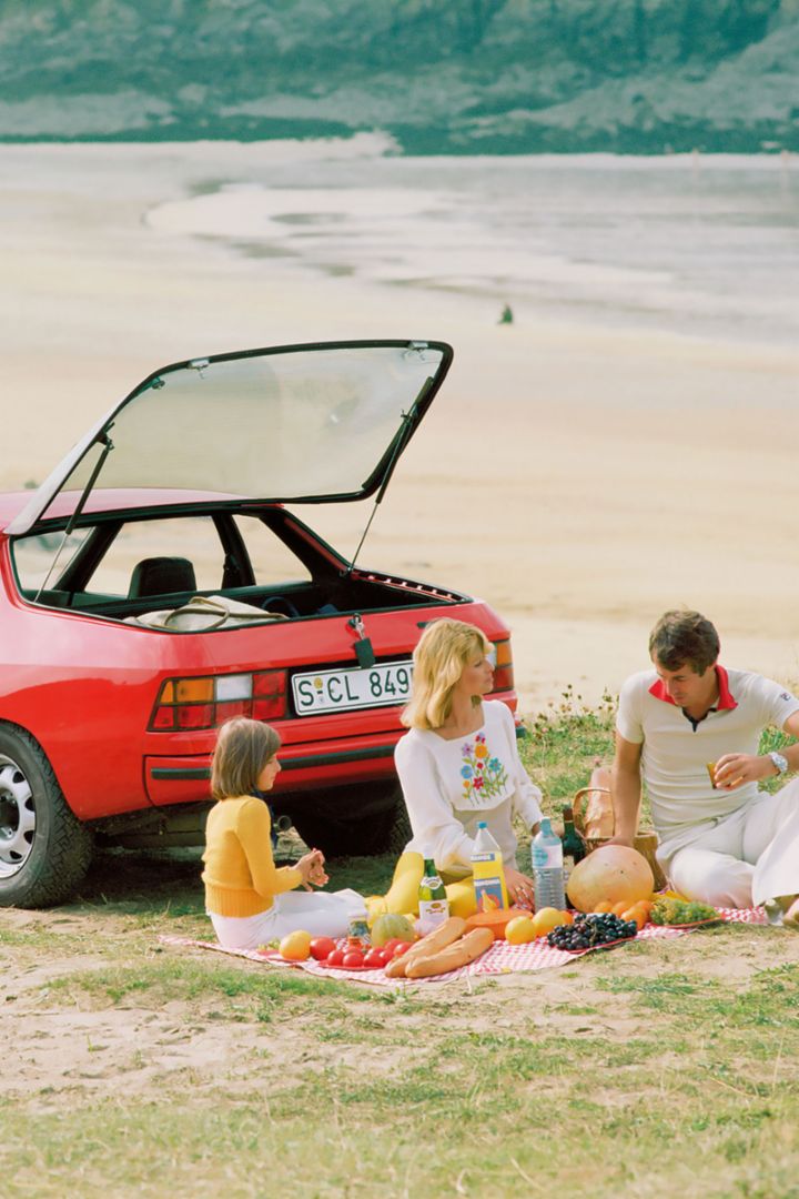 Eine Familie macht ein Picknick vor einem roten Porsche 924 am Strand, mit einem weiten Blick auf das Meer im Hintergrund.