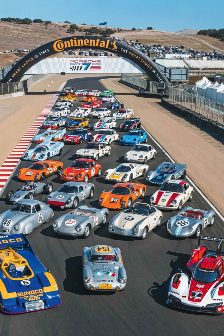 Multiple historic and modern race cars are lined up on a racetrack beneath a bridge displaying the Continental logo.