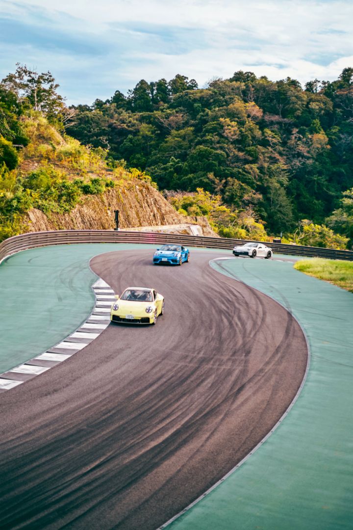 Three Porsche vehicles drive through a sweeping corner on a racetrack surrounded by trees.