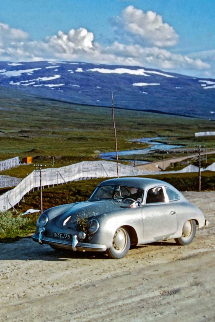 A classic Porsche is parked on a gravel road in front of a wide mountain landscape with patches of snow.