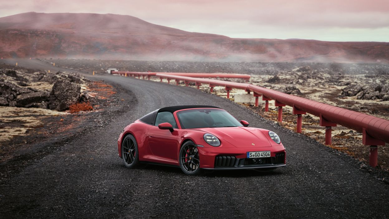 A red Porsche 911 is parked on a winding, dusty road with volcanic terrain surrounding it and a bright sky above.
