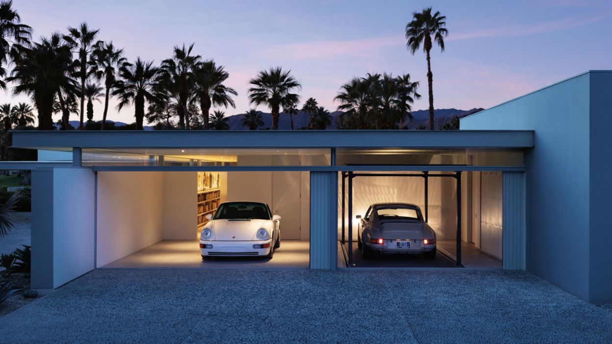 Two classic Porsches stand in a modern, illuminated garage building, surrounded by palm trees and at dusk.