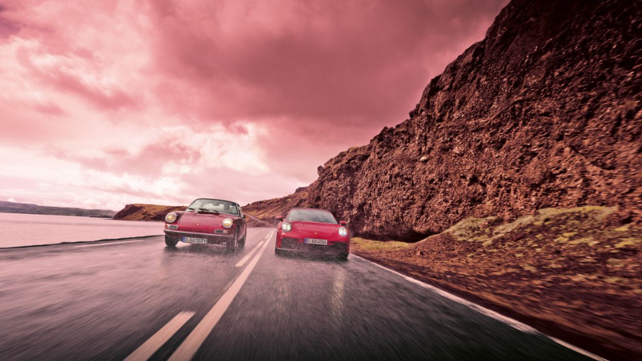 Two Porsche cars drive side by side along a wet road by a rocky cliff with a dramatic, stormy sky above. 