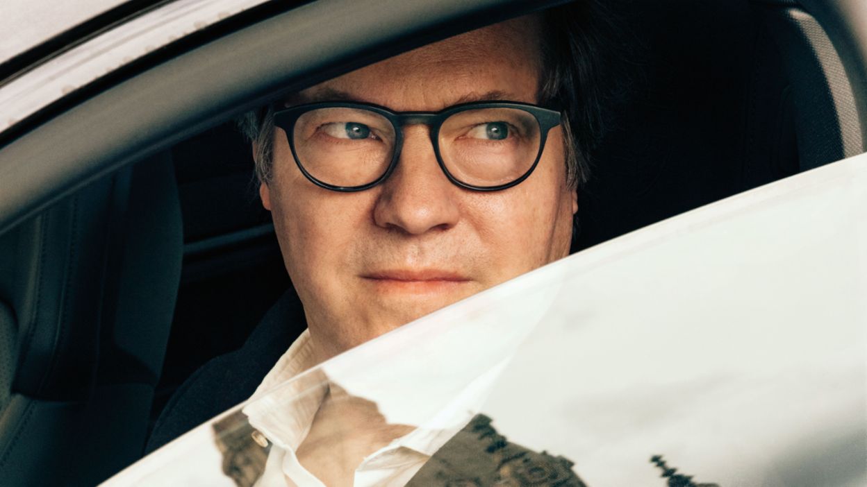 A man wearing glasses looks thoughtfully out of a car window, while a historic building is reflected in the window pane.