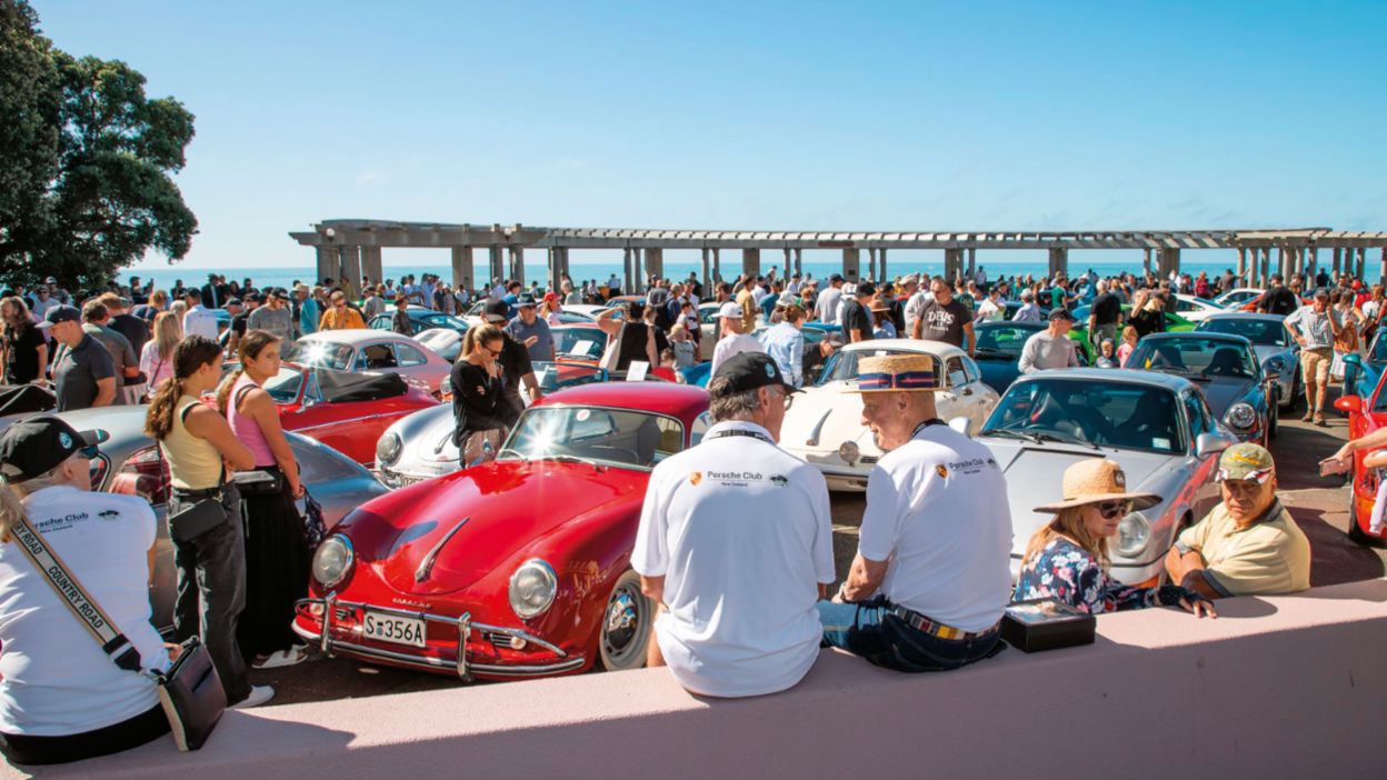 A group of people are looking at sports cars from different eras parked by the sea. 