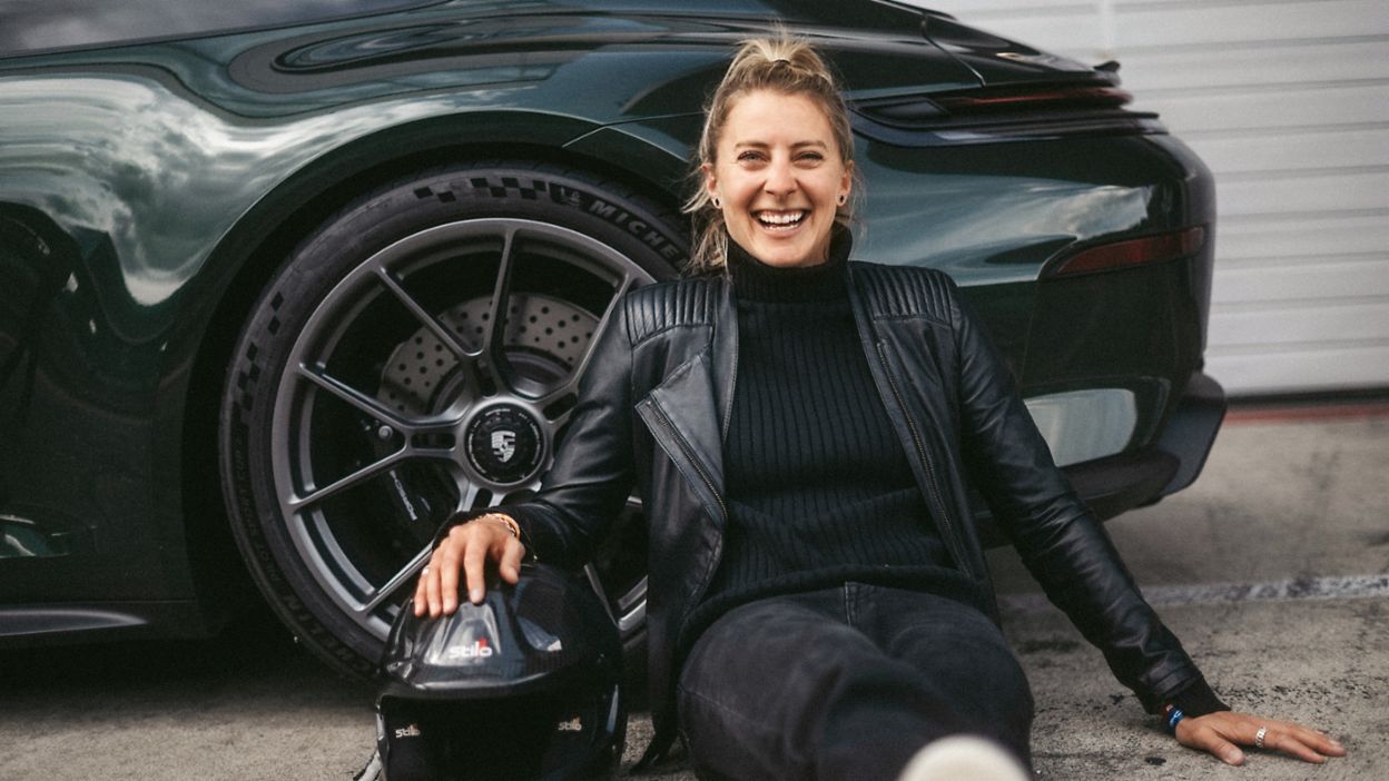 A smiling woman sits on the ground next to a green Porsche 911, holding her helmet in her hand.