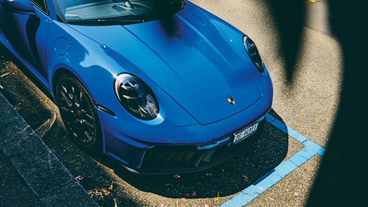 Ein blauer Porsche-Sportwagen steht in einer markierten Parklücke, teilweise im Schatten und mit Blick auf die Fahrzeugfront.