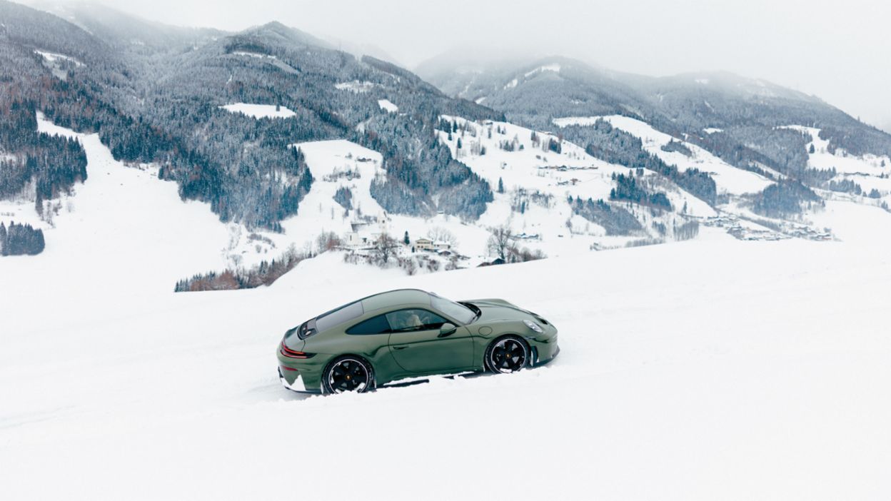 Ein Porsche 911 fährt im Schnee, während die winterliche Berglandschaft im Hintergrund zu sehen ist.