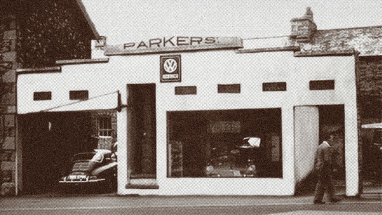 A historic photograph shows a small car dealership with vehicles outside and inside the showroom.