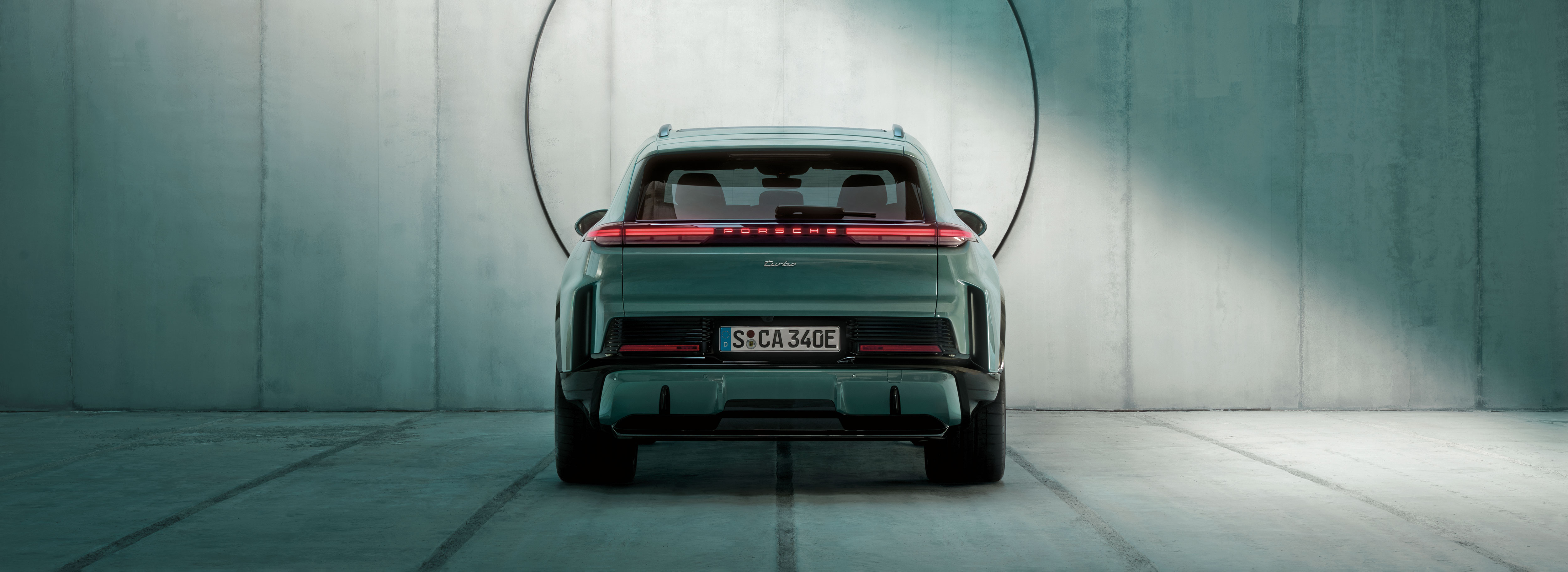 Heckansicht eines grünen Porsche Cayenne Turbo, der frontal vor einer puristischen Betonwand steht, in die ein Kreis eingelassen ist. 